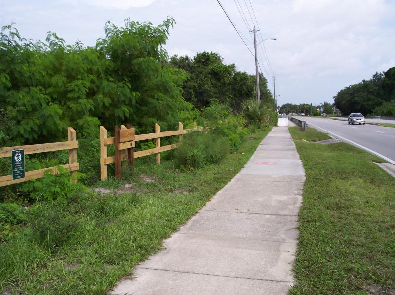A concrete sidewalk bordered by green grass and shrubs, with a wooden fence along one side. The scene includes a grassy area with a sign at the fence, and a road visible on the right side with a passing car. The sky is overcast, suggesting a cloudy day. Turkey Creek mountain bike trail.