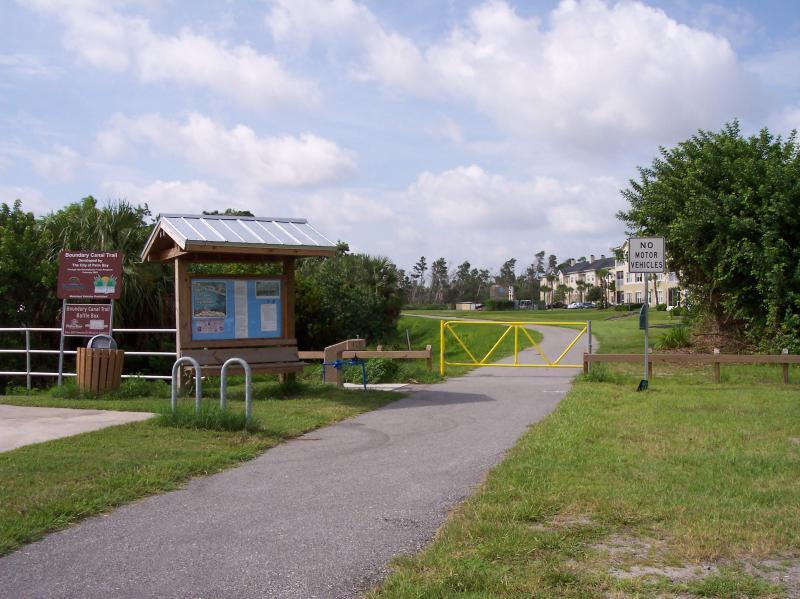 A pathway leading to a trail entrance with a wooden signpost displaying information about the Discovery Canal Trail. There is a gated entrance ahead, and a "No Motor Vehicles" sign nearby. Lush greenery surrounds the area, with residential buildings visible in the background under a partly cloudy sky. Turkey Creek mountain bike trail.