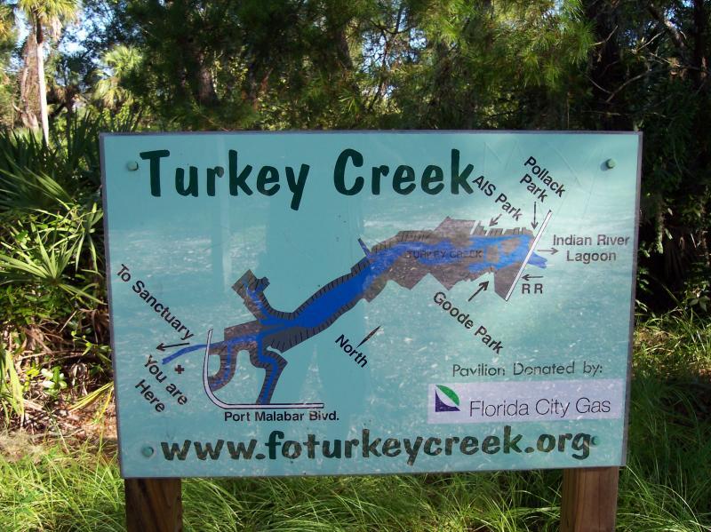 Sign for Turkey Creek showcasing a map of the area, including marked locations such as AIS Park, Pollack Park, Goode Park, and the Indian River Lagoon. The sign includes directional indicators and a web address for more information. Surrounding vegetation is visible in the background. Turkey Creek mountain bike trail.