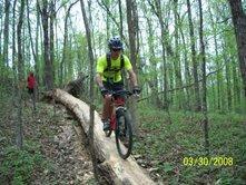 A person riding a mountain bike on a fallen log in a forested area, with green trees and foliage surrounding the trail. The Ridgeland Trails mountain bike trail.