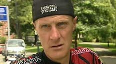 A man wearing a black cap and a red cycling jersey stands outdoors, with trees and a road sign in the background. He has a serious expression on his face.