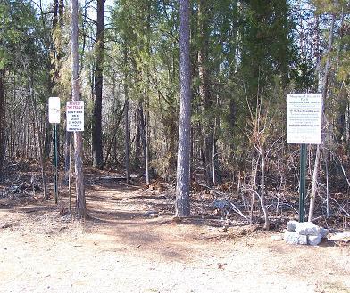 A dirt path leading into a wooded area, flanked by two signs on metal posts. The signs contain text, likely providing information or rules about the area. Surrounding trees and underbrush indicate a natural setting. Sherman Branch Park Mtb Trail mountain bike trail.