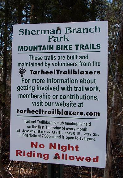 A sign in Sherman Branch Park displaying information about mountain bike trails maintained by volunteers from Tarheel Trailblazers. The sign includes details on how to get involved with trailwork and mentions that night riding is not allowed. There is also information about the monthly club meeting at Jack's Bar & Grill in Charlotte. Sherman Branch Park Mtb Trail mountain bike trail.