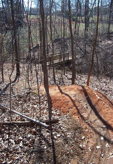 A dirt jump ramp is visible in a wooded area, surrounded by bare trees and fallen leaves. In the background, a wooden bridge can be seen crossing a small area of grass. The scene is set on a sunny day, showcasing a peaceful outdoor environment. Sherman Branch Park Mtb Trail mountain bike trail.