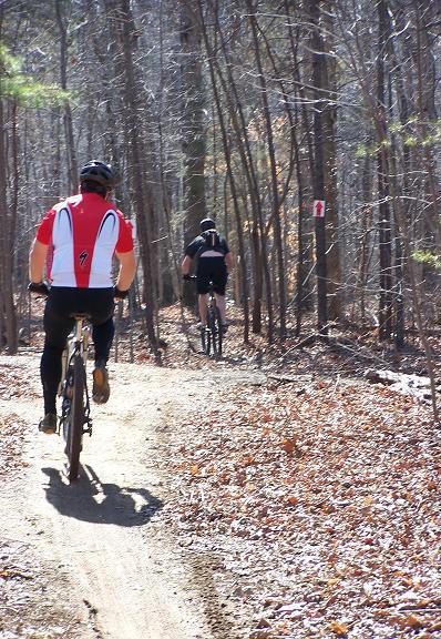 Two mountain bikers riding along a dirt trail in a wooded area during a sunny day. One biker is wearing a red and white jersey, while the other is dressed in black. The trail is surrounded by trees, and there are fallen leaves on the ground. A trail marker is visible in the background. Sherman Branch Park Mtb Trail mountain bike trail.