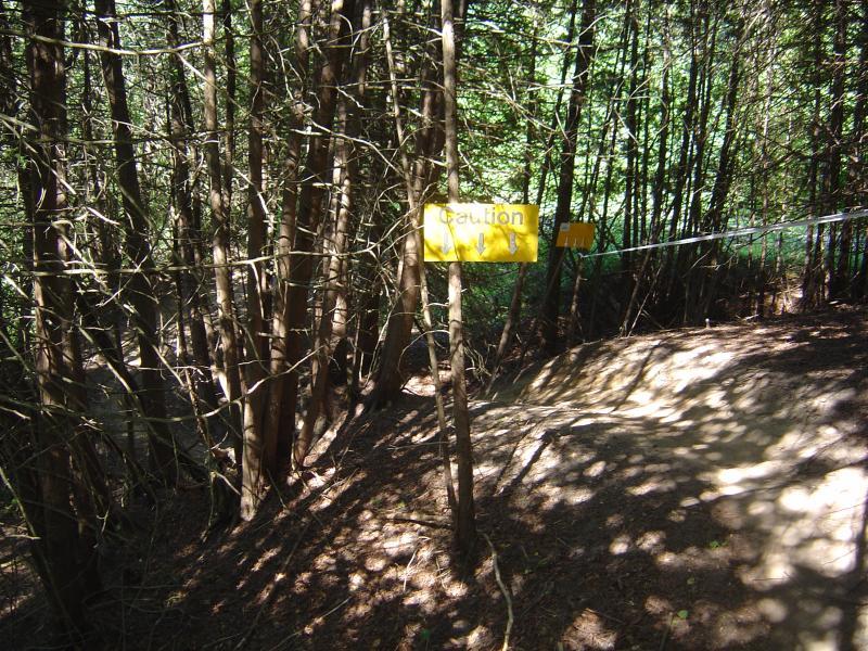 A wooded area featuring a caution sign in a bright yellow color. The sign is partially obscured by branches and is positioned near a trail leading down a slope. Sunlight filters through the trees, casting shadows on the ground. Albion Hills mountain bike trail.