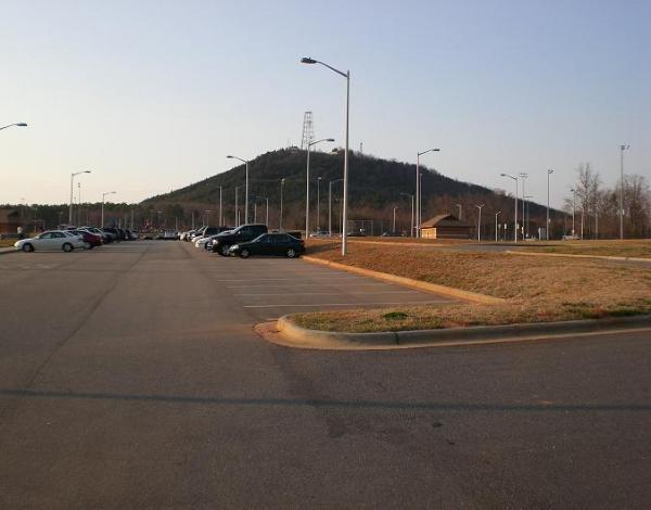 A wide view of a parking lot lined with cars, leading towards a distant hill with a communication tower at the top. The scene is set during the late afternoon, with a clear sky and sparse trees in the background. George Poston Park mountain bike trail.