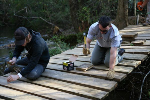 Two individuals are working together on a wooden pathway over a stream. One person is kneeling and using a hammer, while the other is seated, tying something with a rope. They are surrounded by trees and nature, indicating an outdoor setting that suggests a community service or construction project. George Poston Park mountain bike trail.