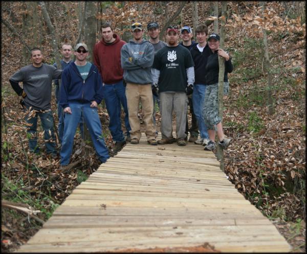 A group of ten men standing on a newly constructed wooden bridge in a wooded area, surrounded by fallen leaves and trees. The men are dressed in casual outdoor clothing and are smiling at the camera. George Poston Park mountain bike trail.