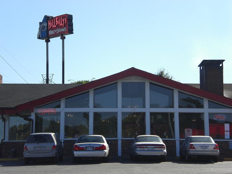 A restaurant exterior featuring large windows, a red roof, and a sign that reads "Hillbilly's BBQ." The building has a welcoming "OPEN" sign, with several parked cars in front. George Poston Park mountain bike trail.