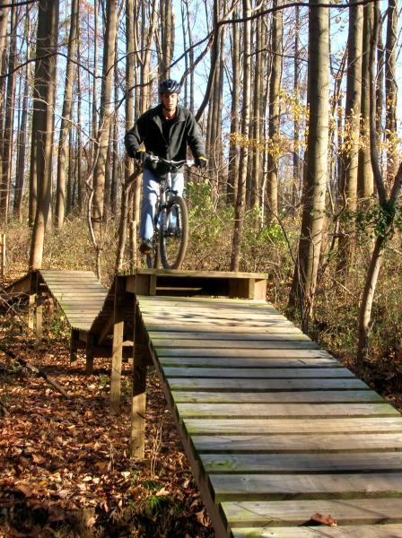 A person riding a mountain bike over a wooden ramp in a forested area. The cyclist is wearing a helmet and appears to be in mid-jump on the ramp, surrounded by trees with autumn leaves on the ground. Middle Run Natural Area mountain bike trail.