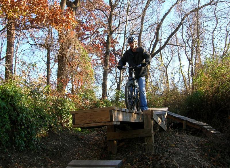 A person in a black jacket and helmet is riding a mountain bike on a wooden ramp in a wooded area during autumn. The background features trees with colorful leaves and underbrush, indicating a natural outdoor setting. Middle Run Natural Area mountain bike trail.