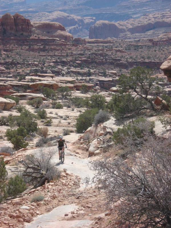 A mountain biker rides along a dirt trail winding through a rugged desert landscape, featuring rocky formations, sparse vegetation, and distant mountains under a clear blue sky. Gold Bar Rim mountain bike trail.