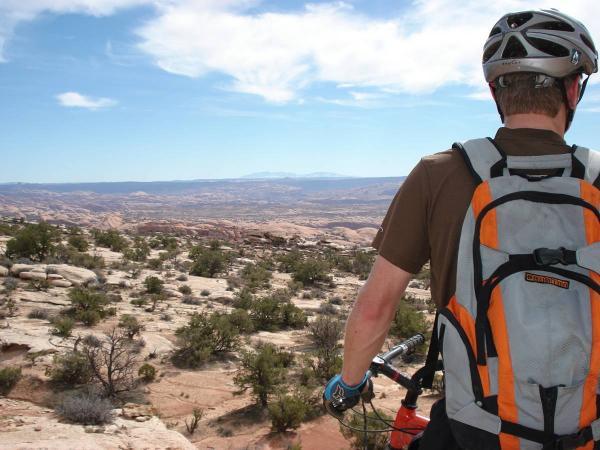 A person in a brown shirt and a gray backpack stands on a rocky outcrop, looking out over a vast desert landscape under a blue sky with scattered clouds. The view includes distant mountains and vegetation scattered across the terrain. The individual is wearing a helmet and holding a mountain bike, suggesting they are enjoying outdoor recreation. Gold Bar Rim mountain bike trail.