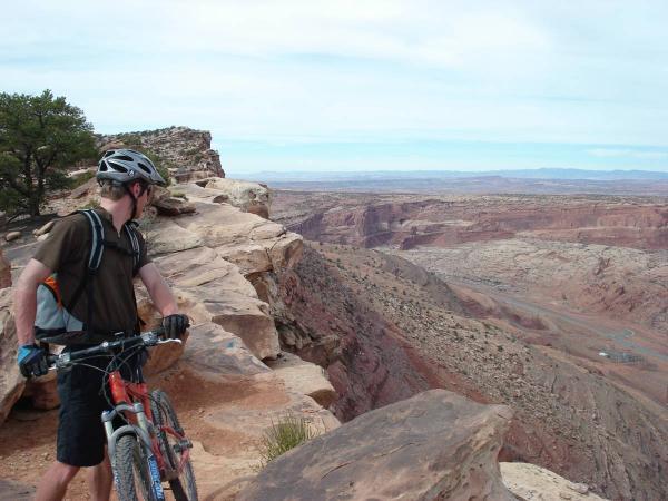 A person stands on the edge of a rocky cliff, looking out over a vast desert landscape. They are wearing a helmet and a brown shirt, and have a mountain bike beside them. The scenery features layered red and brown rock formations, with a clear sky above. Gold Bar Rim mountain bike trail.
