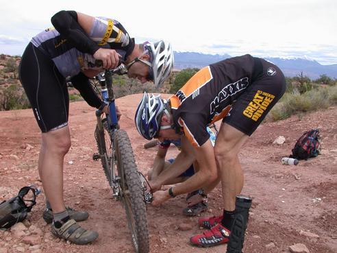 Three mountain bikers working together to fix a flat tire on a rocky trail. One biker is kneeling beside the bike, inspecting the wheel, while the other two are focusing on the repair. The landscape is rugged, with distant mountains visible in the background. Gold Bar Rim mountain bike trail.