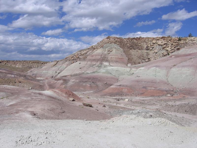 A landscape featuring layered hills with hues of pink, green, and beige, under a partially cloudy blue sky. The foreground shows a mix of light-colored soil and rocks, while the hills in the background have a more rugged, rocky appearance. Lunch Loops mountain bike trail.