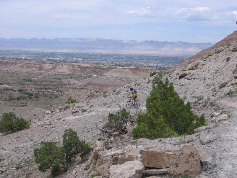 A mountain biker rides along a rocky trail on a hillside, with a vast landscape of rolling hills and distant mountains visible in the background under a partly cloudy sky. Lunch Loops mountain bike trail.