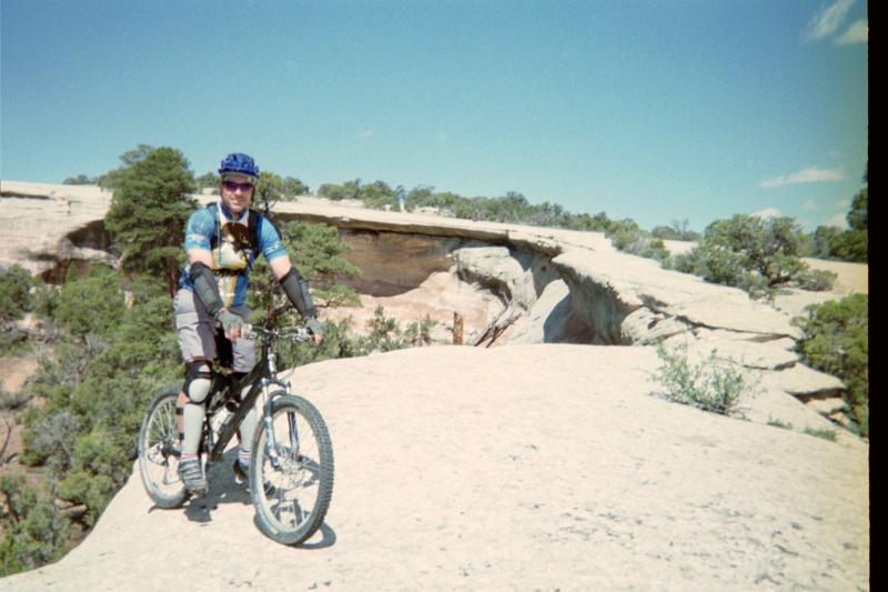 A mountain biker wearing a blue helmet and protective gear poses next to his bike on a rocky outcrop. In the background, trees and a cliff formation are visible under a clear blue sky. Lunch Loops mountain bike trail.