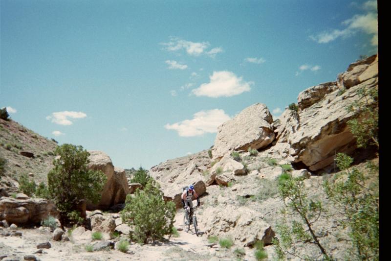 A person riding a mountain bike along a narrow trail surrounded by large boulders and greenery, with a bright blue sky and scattered clouds above. Lunch Loops mountain bike trail.