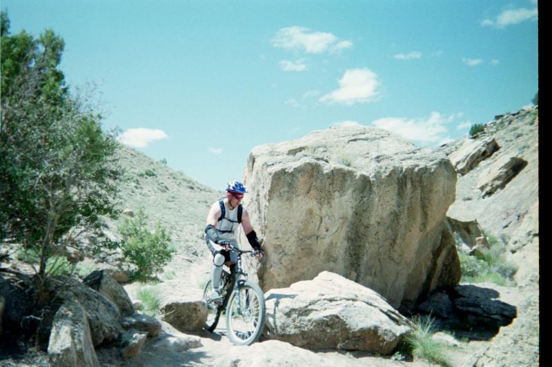 A mountain biker navigating a rocky trail with a large boulder nearby, surrounded by desert terrain and blue skies with scattered clouds. Lunch Loops mountain bike trail.