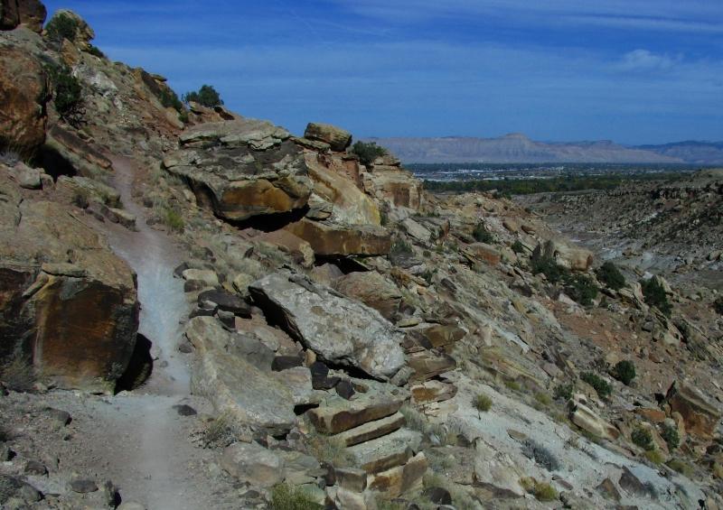 A rocky hillside with a dirt path winding through it, leading to a view of distant hills and a valley below under a clear blue sky. Lunch Loops mountain bike trail.