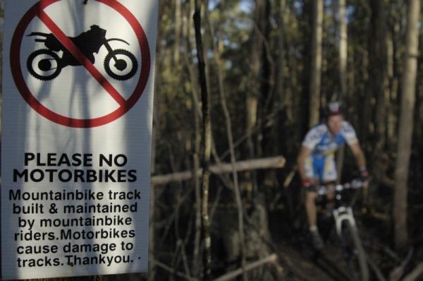 A mountain biker riding on a trail surrounded by trees, with a nearby sign that reads "Please No Motorbikes" featuring a motorcycle icon and a message about the impact of motorbikes on mountain bike tracks. Wombat Track mountain bike trail.