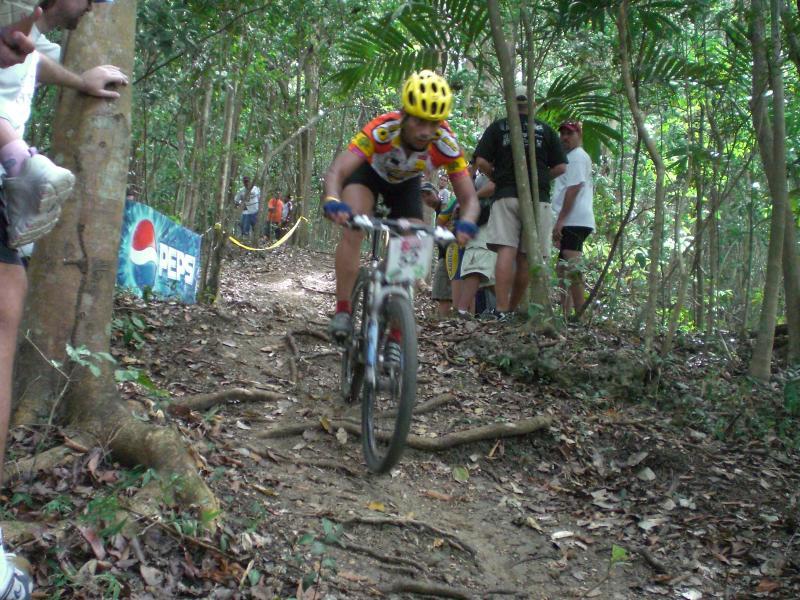 A mountain biker in a yellow helmet navigates a rough, root-covered trail through a lush forest, while spectators watch from the side, some near a Pepsi banner. Monagas mountain bike trail.