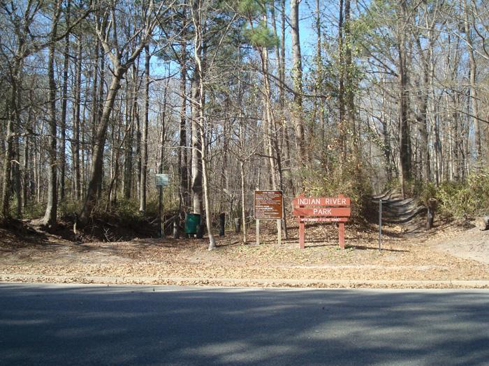 Entrance to Indian River Park, featuring wooden signs and a natural, wooded environment with trees and a path leading into the park. The scene is set along a road with fallen leaves on the ground. Indian River Park mountain bike trail.