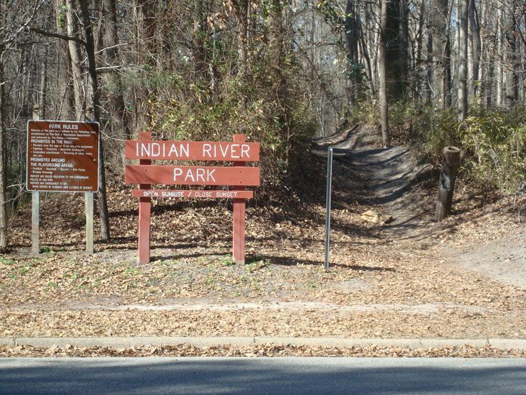 Sign marking the entrance to Indian River Park, featuring park rules on a brown informational sign to the left, with a wooded trail leading into the park in the background. The area is surrounded by trees and scattered leaves. Indian River Park mountain bike trail.