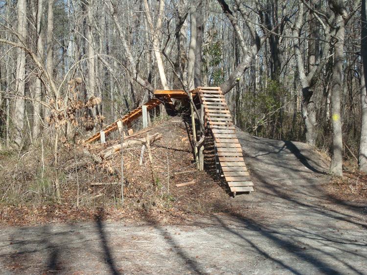 A wooden ramp built into a small hill in a wooded area, surrounded by bare trees. The ramp is made of planks and leads up from a dirt path, suggesting a setup for biking or outdoor activities. Indian River Park mountain bike trail.