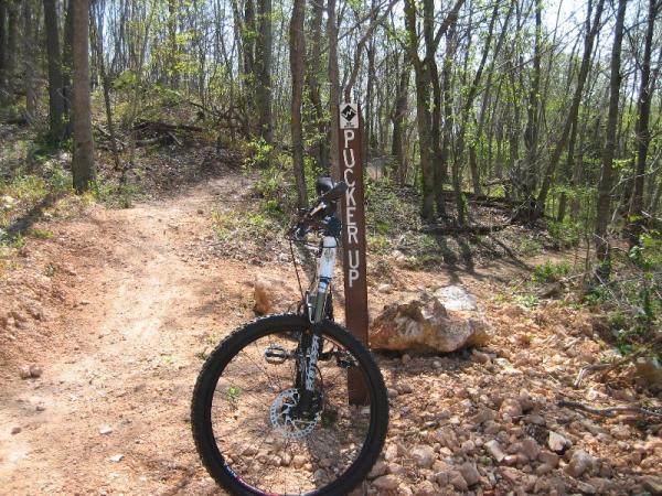 A mountain bike leaning against a trail sign that reads "PUCKER UP," surrounded by a wooded area and rough terrain. The ground is gravelly with scattered rocks, and lush greenery can be seen in the background, indicating a natural outdoor setting. Slaughter Pen Trail mountain bike trail.