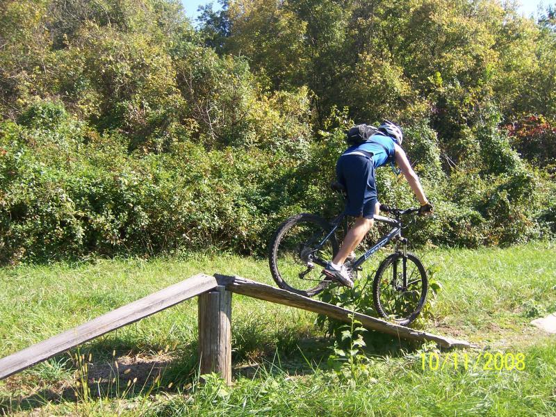 A mountain biker navigating a wooden ramp in a lush, green outdoor setting, showcasing a dynamic moment of balance and skill as the bike ascends the structure. Slaughter Pen Trail mountain bike trail.