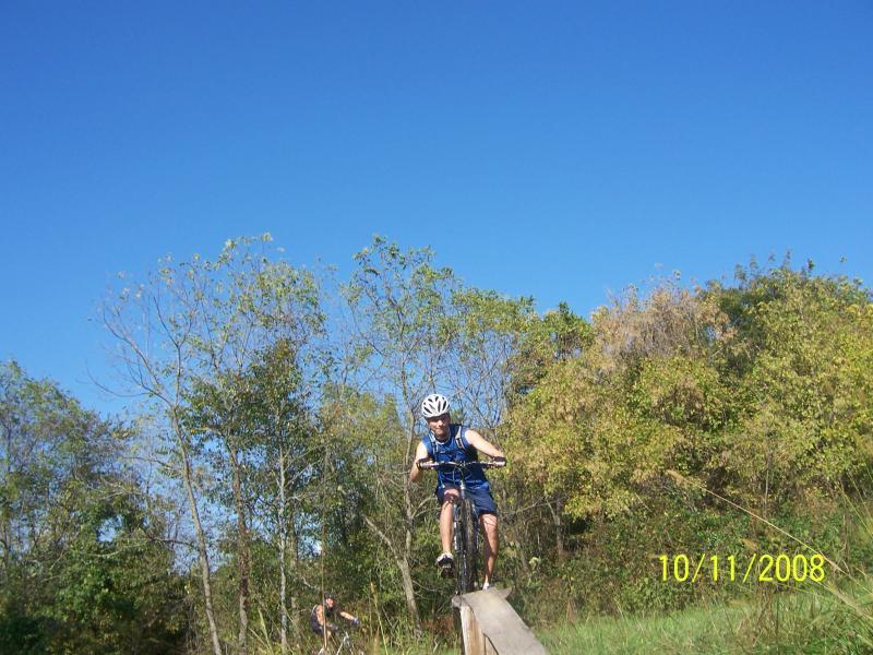 A young cyclist in a helmet performing a stunt by jumping off a wooden ramp on a sunny day. Lush greenery and trees can be seen in the background under a clear blue sky. Slaughter Pen Trail mountain bike trail.