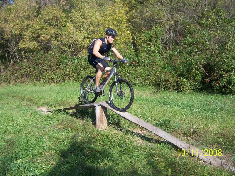 A mountain biker riding on a wooden ramp in a grassy area, surrounded by trees. The cyclist is wearing a helmet and a sleeveless shirt, demonstrating balance and skill as he navigates the trail. Slaughter Pen Trail mountain bike trail.
