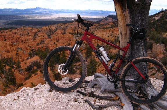 A red mountain bike is positioned on a rocky ledge overlooking a vast canyon landscape. The scene features layered rock formations in shades of orange and brown, with trees scattered throughout. In the background, the horizon extends into distant blue mountains under a cloudy sky. Thunder Mountain mountain bike trail.