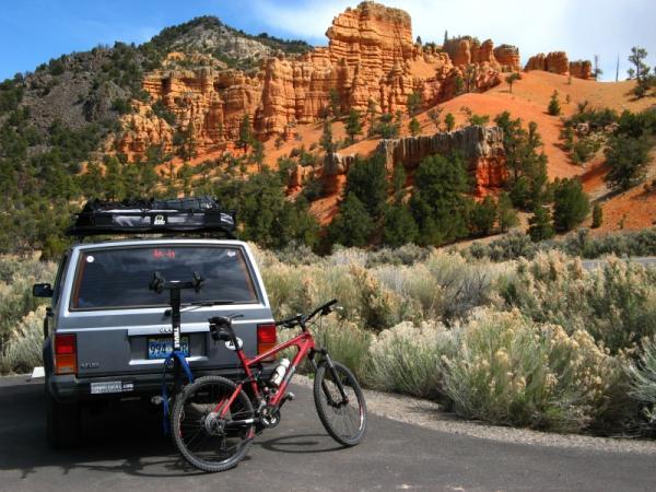 A parked gray SUV with mountain bikes mounted on the back, set against a backdrop of orange rock formations and lush greenery under a partly cloudy sky. Thunder Mountain mountain bike trail.