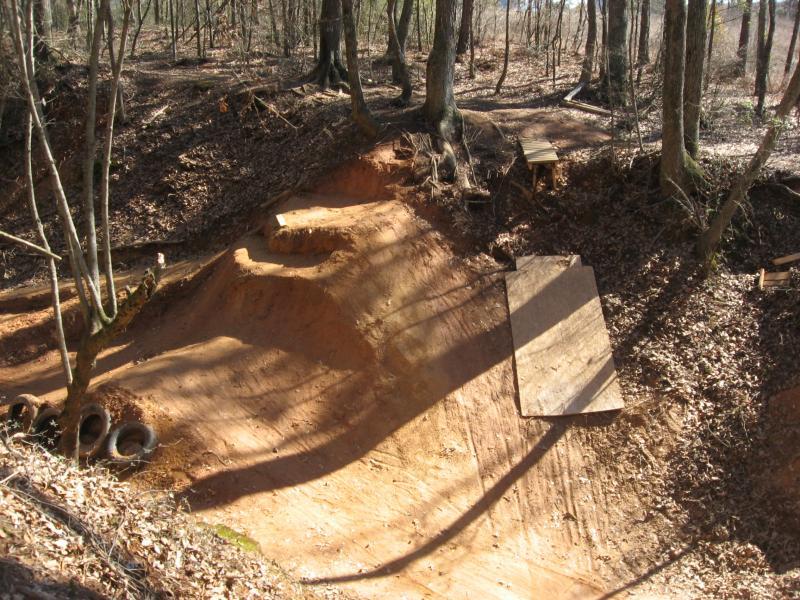 A dirt bike jump and ramp area in a woodland setting, surrounded by trees and leaf-covered ground. The jump features a sandy surface with various elevations and wooden planks serving as launch points. 441 Trails mountain bike trail.