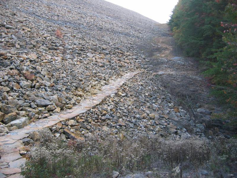A rocky hillside with a narrow stone pathway winding through it, surrounded by sparse vegetation and trees on one side. The path leads upward, indicating a hiking trail in a natural setting. Raccoon Mountain Trail Network mountain bike trail.