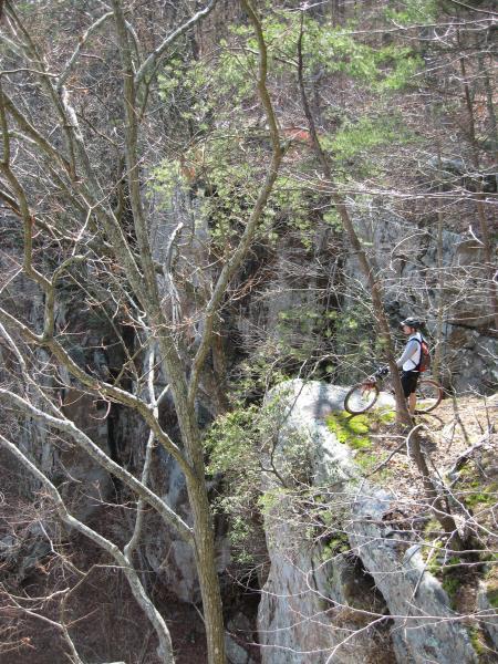 A cyclist in a white shirt and black helmet stands near the edge of a rocky cliff, with a mountain bike beside them. The scene is surrounded by bare trees and patches of greenery, showcasing a natural landscape in early spring. Raccoon Mountain Trail Network mountain bike trail.