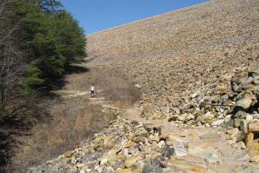 A rocky landscape with a steep earthen dam in the background. In the foreground, a path made of stones meanders through sparse vegetation. On the left side, there are clusters of trees. A person is walking along the path, providing a sense of scale to the vast terrain. The sky is clear and blue. Raccoon Mountain Trail Network mountain bike trail.