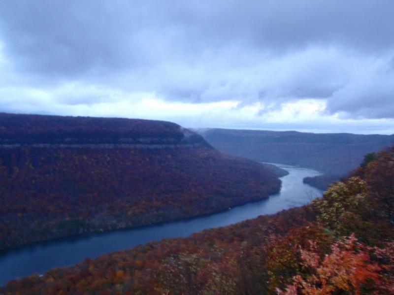A scenic view of a winding river surrounded by vibrant autumn foliage, with rolling hills and a cloudy sky in the background. The landscape features a dramatic cliff on the left side, showcasing the colorful fall leaves. Raccoon Mountain Trail Network mountain bike trail.