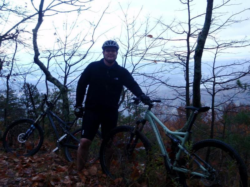 A mountain biker stands in a wooded area, surrounded by trees and autumn foliage, with two mountain bikes beside him. He is wearing a helmet and a black jacket, and the scene is set on a cloudy day with a view of the distant landscape. Raccoon Mountain Trail Network mountain bike trail.