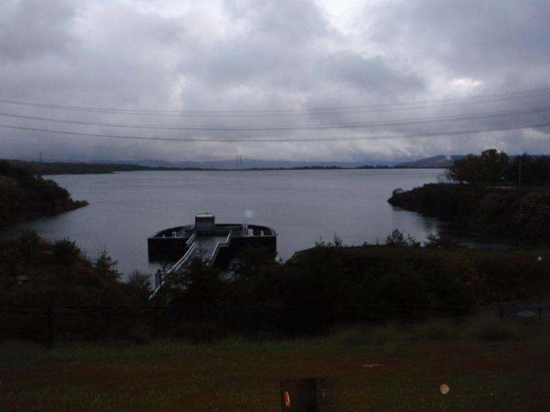 A calm body of water surrounded by grassy banks and trees, with a dock or platform extending into the water. The sky is overcast, creating a muted, gray atmosphere. In the distance, faint outlines of hills and power lines can be seen against the cloudy backdrop. Raccoon Mountain Trail Network mountain bike trail.