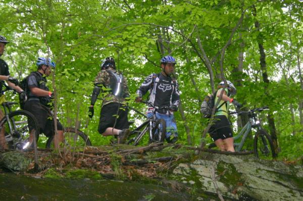 A group of mountain bikers in cycling gear are navigating a rocky path through a lush, green forest. They are positioned among trees, with some riders dismounting their bikes while others are adjusting their gear. The scene captures the essence of outdoor adventure and teamwork in a natural setting. Blue Mountain Reservation mountain bike trail.