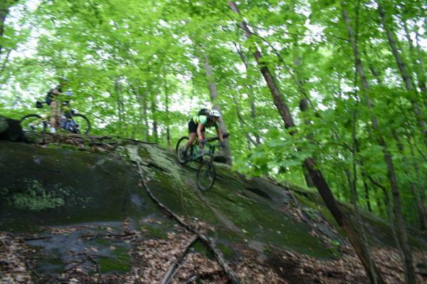 Mountain biker descending a rocky slope surrounded by lush green trees and foliage, with another cyclist in the background. Blue Mountain Reservation mountain bike trail.