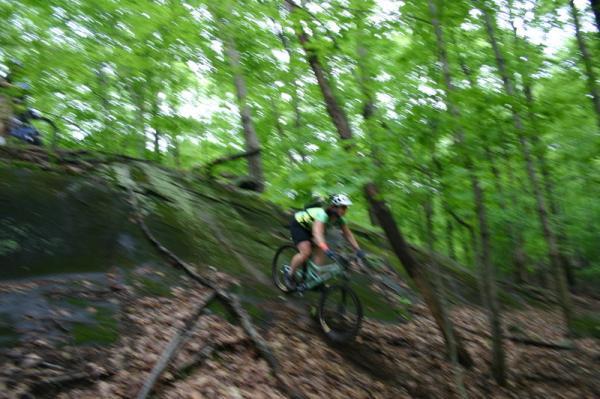 A mountain biker navigating a steep, rocky trail in a lush green forest, with trees surrounding the path and fallen leaves covering the ground. The image captures motion, suggesting an energetic descent. Blue Mountain Reservation mountain bike trail.