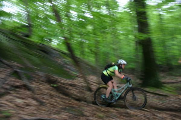 A mountain biker navigating a winding path through a lush green forest, captured in motion with a blurred background that conveys speed and excitement. The rider is wearing a helmet and sporty attire, enhancing the dynamic nature of the scene. Blue Mountain Reservation mountain bike trail.