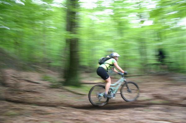 A person riding a mountain bike along a winding trail in a lush green forest, captured in motion with a blurred background, conveying speed and energy. The cyclist is wearing a helmet and a bright yellow top, surrounded by tall trees and dense foliage. Blue Mountain Reservation mountain bike trail.
