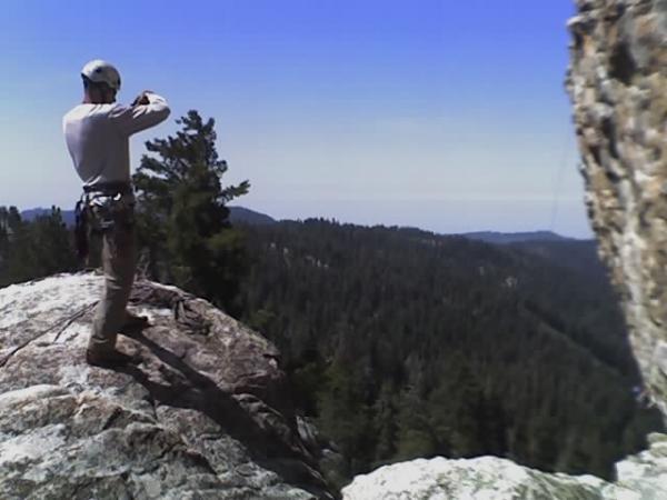 A person wearing a climbing helmet and gear stands on a rocky outcrop, overlooking a vast forested landscape under a clear blue sky. The individual appears to be taking a photograph or enjoying the view. Buck Rock mountain bike trail.